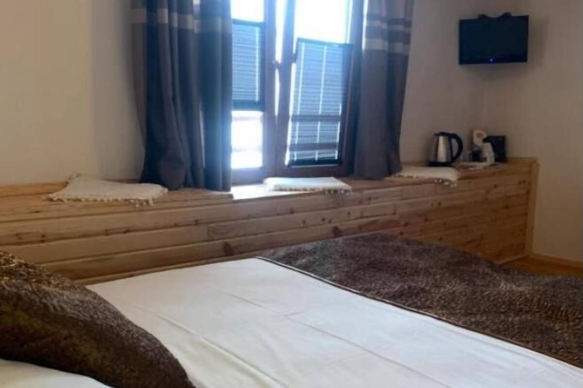 Une pièce avec un lit, une fenêtre avec des rideaux, un banc en bois et un téléviseur fixé au mur.