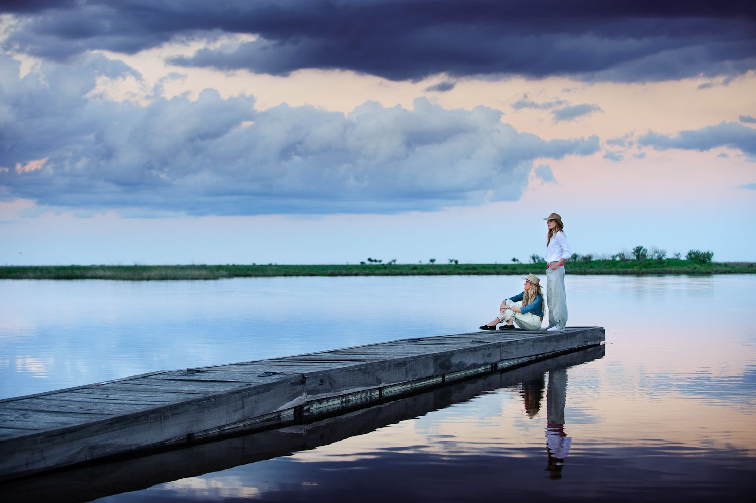 Une jetée en bois s’étendant sur une étendue d’eau calme, avec une personne debout et une autre assise à son extrémité.