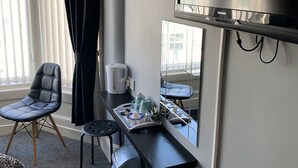 Et hotelværelse med et stort fladskærms-tv på væggen, en sort læderstol, et lille bord med en bakke og diverse genstande, en kaffemaskine og en skraldespand.