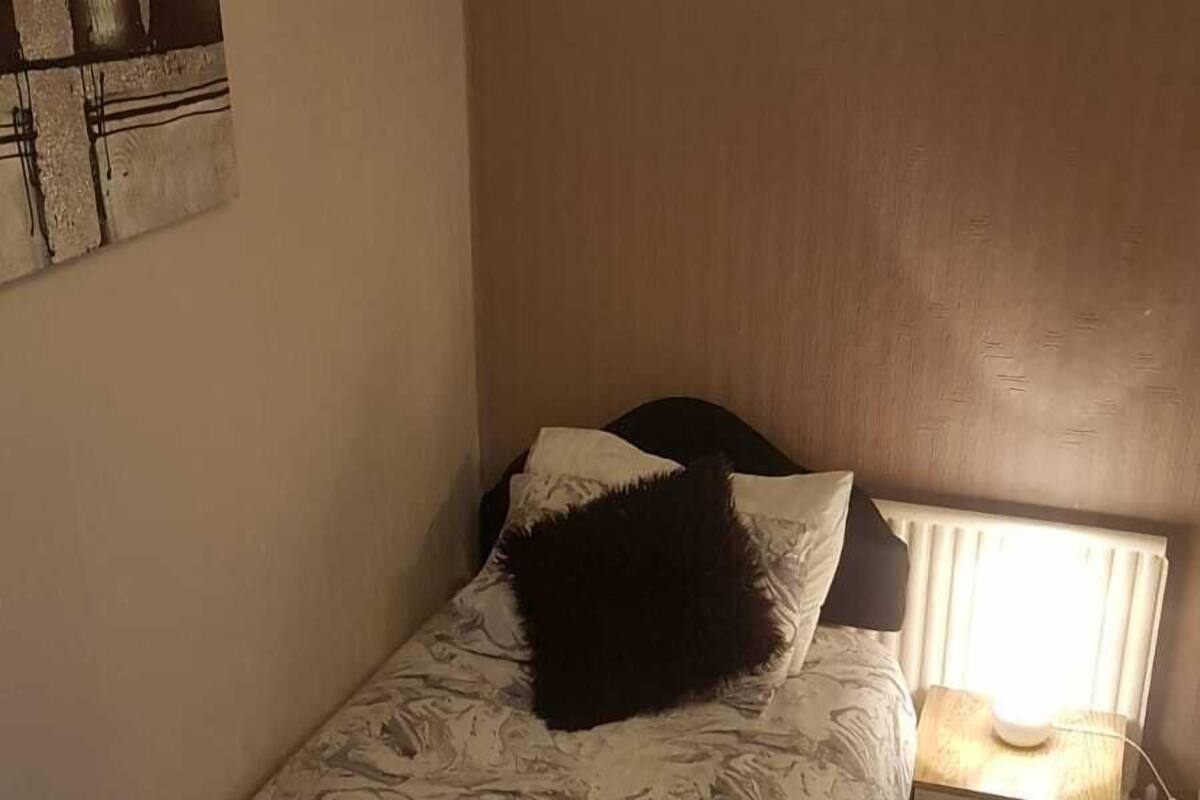 A small, single-bed room with a round ceiling light, a bedside table with a lamp, and a wall-mounted artwork.