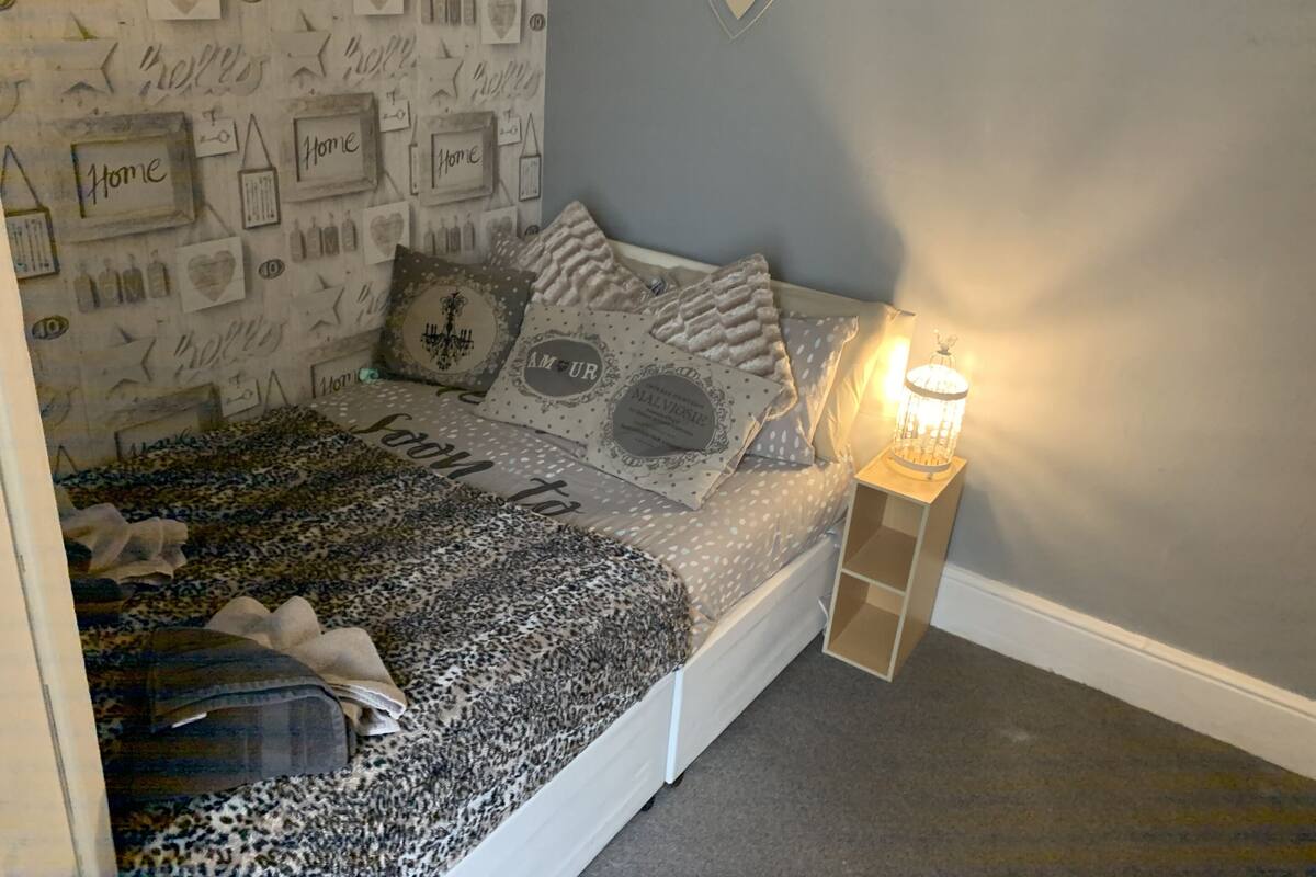 A bedroom with a bed, pillows, a nightstand with a lamp, and a wall decorated with framed items and a heart-shaped sign.