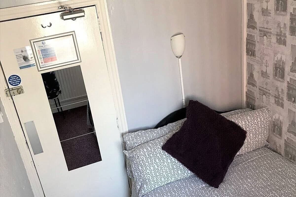 A small room with a bed, a door with a fire safety notice, a mirror, and a wall with a Paris-themed wallpaper.