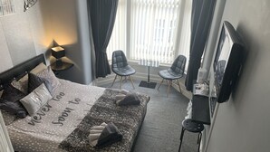 Et hotelværelse med en seng, to stole, et fjernsyn og et vindue med gardiner.