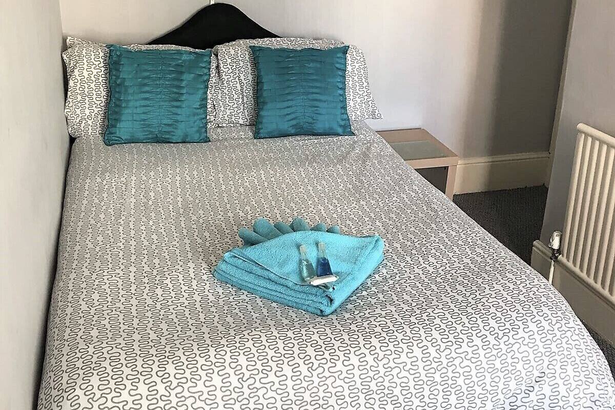 A neatly made bed with patterned bedding and two teal pillows, a folded towel with toiletries, and a bedside table with a lamp.