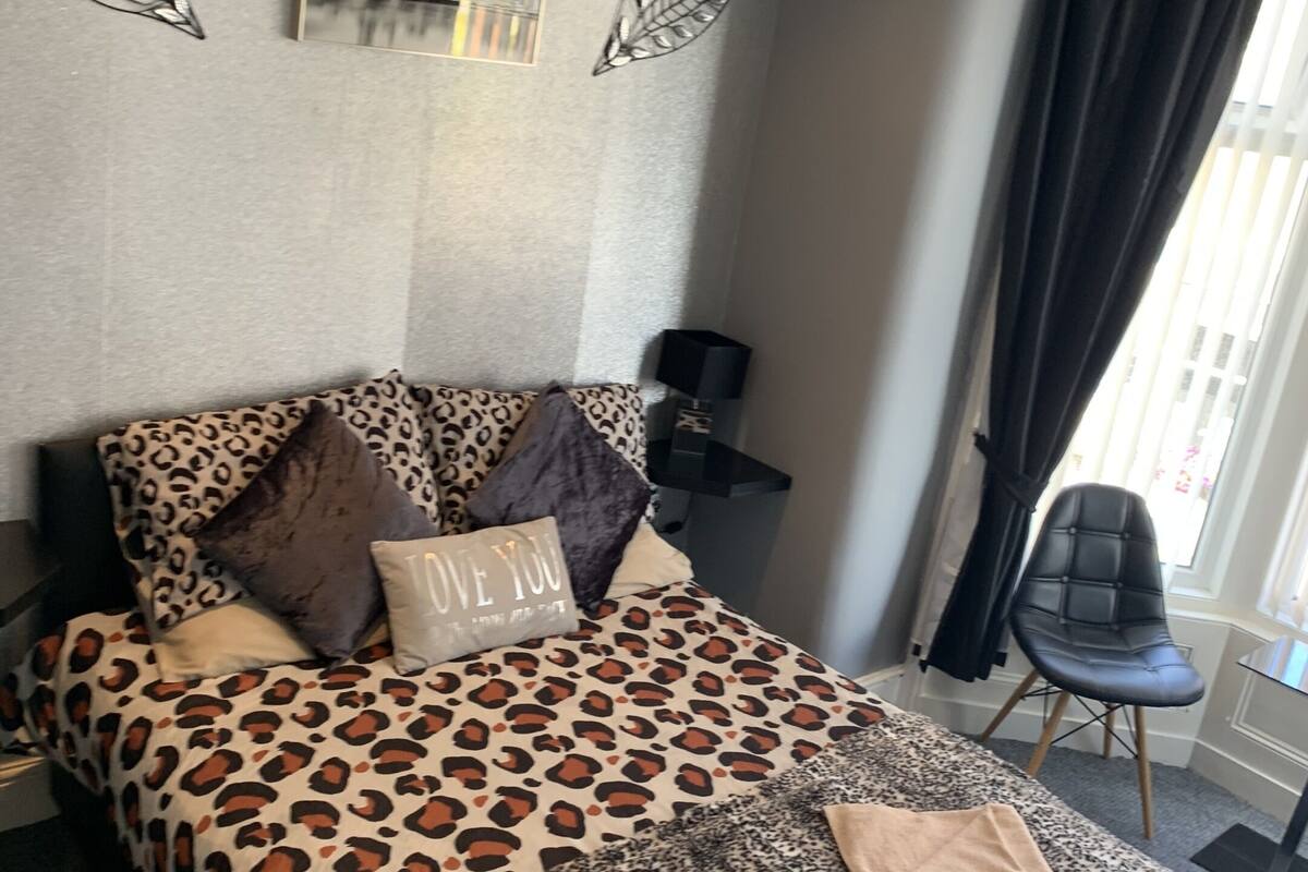 A bedroom with a bed featuring a leopard print bedspread, two pillows, a folded towel, and a decorative wall piece.