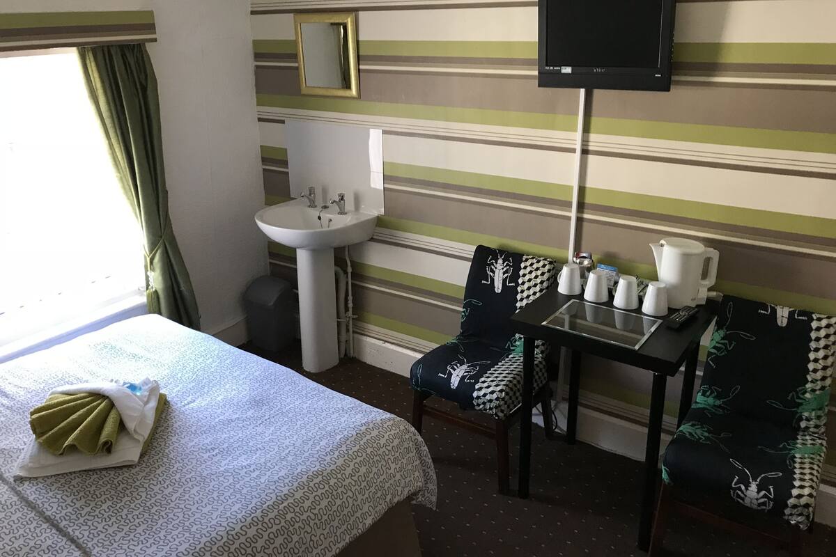A hotel room with a bed, a sink, a table with cups and a kettle, and a TV on the wall.