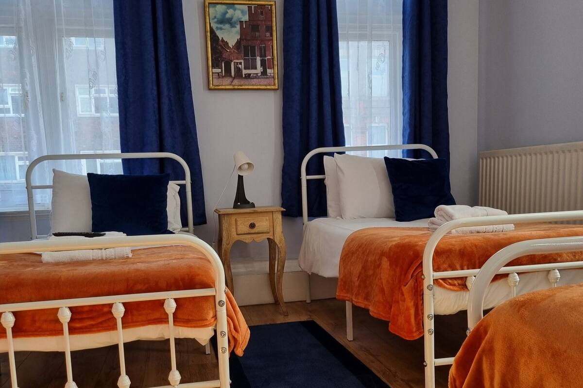 A hotel room with two beds, a small bedside table with a lamp, a blue rug, and a painting on the wall.
