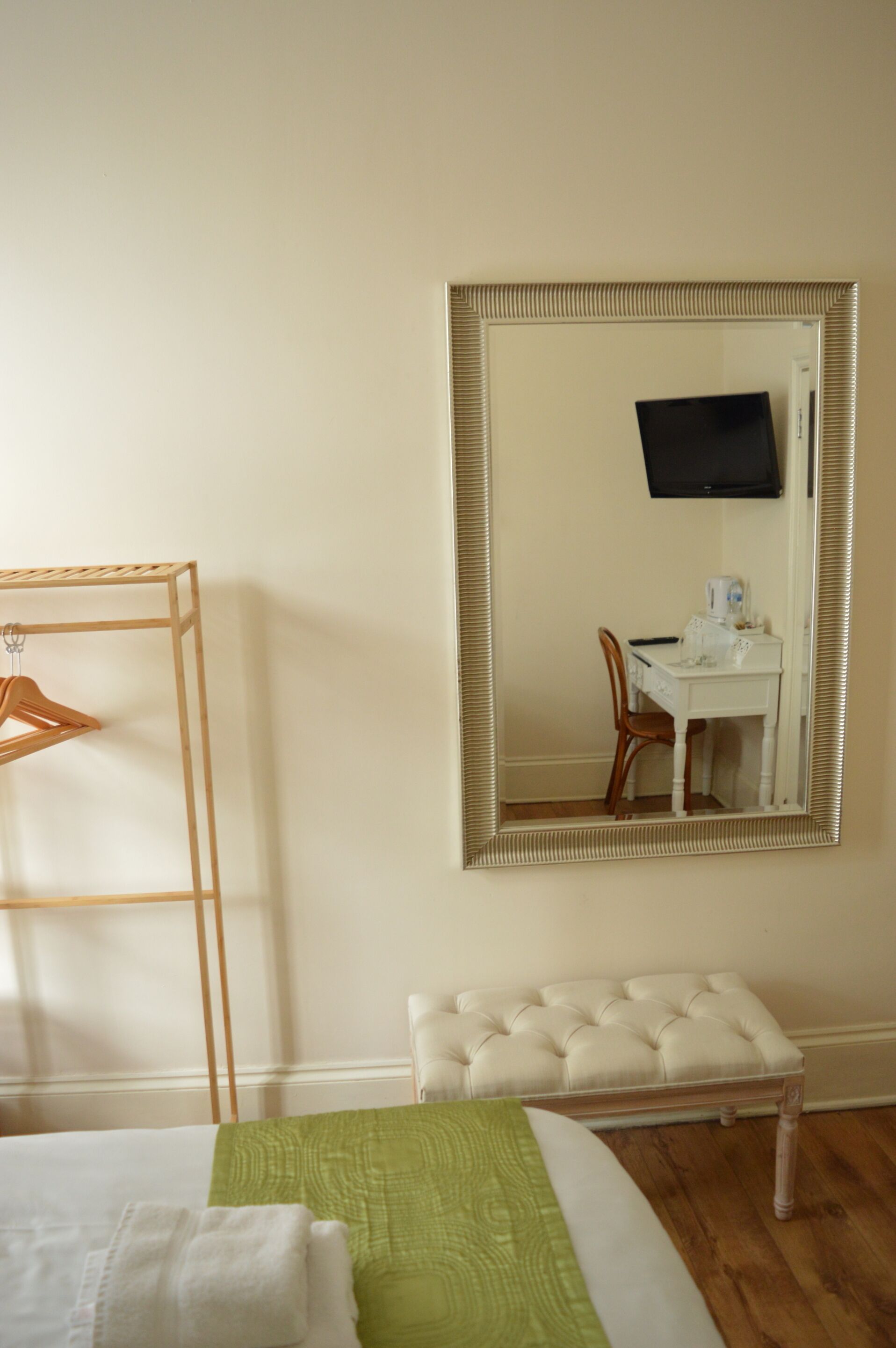 A hotel room with a bed, a green and white bedspread, a wooden hanger rack, a white upholstered bench, a white desk with a chair, and a large mirror with a television above it.