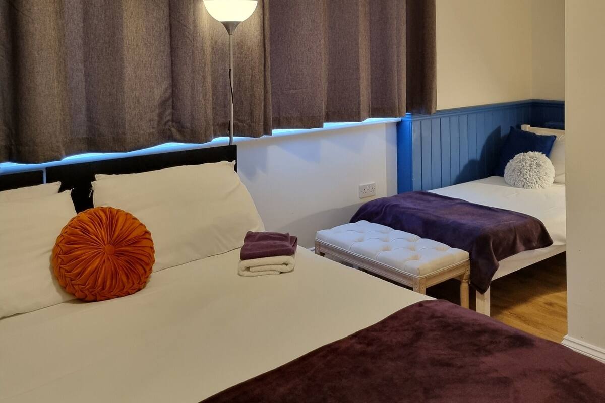 A hotel room with two beds, a nightstand, a lamp, and a large window with curtains.