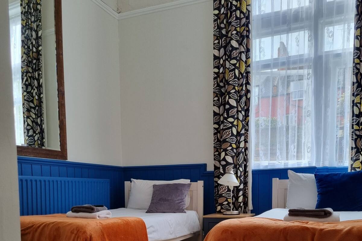 A hotel room with two single beds, a wooden table, and a view of the street through large windows.