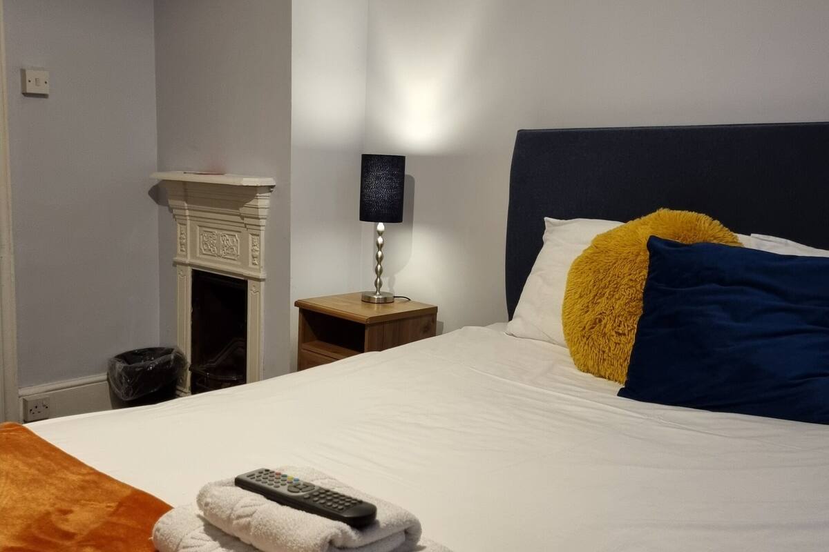 A neatly made bed with a white duvet, a brown throw blanket, and a remote control on top. There is a small wooden nightstand with a lamp beside the bed.