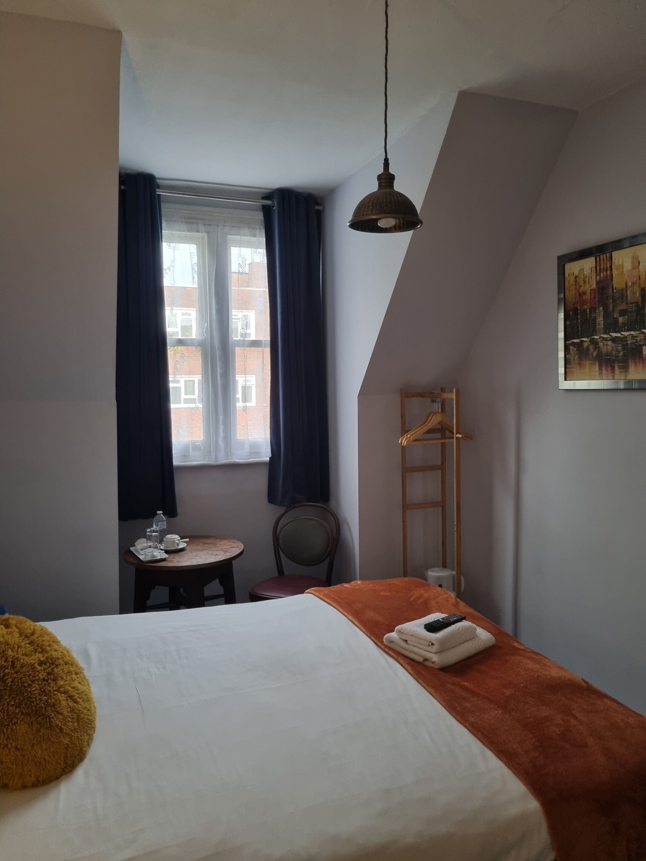 Habitación de hotel con cama, una mesita con silla, ventana con cortinas, lámpara colgante y un cuadro en la pared.