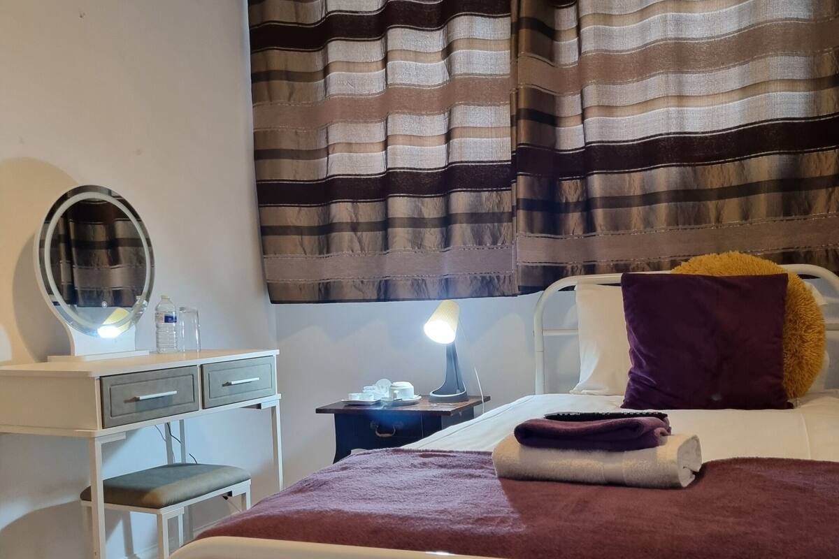 A bedroom with a bed, a bedside table with a lamp, a mirror, and a window with striped curtains.