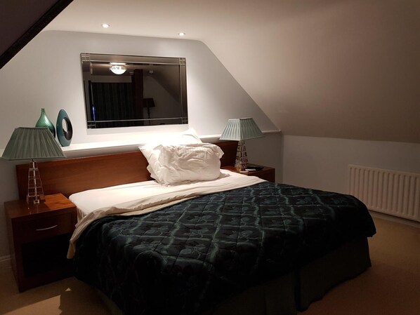 A bedroom with a sloped ceiling, a large bed with a dark quilt, two bedside lamps, a nightstand, a window with a view, and a radiator.