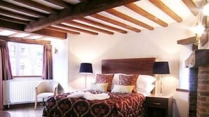 A bedroom with a wooden headboard, a bed with patterned bedding, two bedside lamps, a chair, and a radiator. - Ye Olde Red Lion (Newbury)