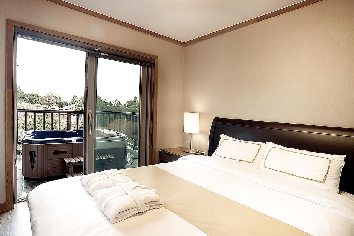 Quarto de hotel com uma cama grande, uma mesa de cabeceira com luminária e uma porta de vidro que dá para uma varanda com hidromassagem.