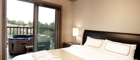 A hotel room with a large bed, a nightstand with a lamp, a sliding glass door leading to a balcony with a hot tub.