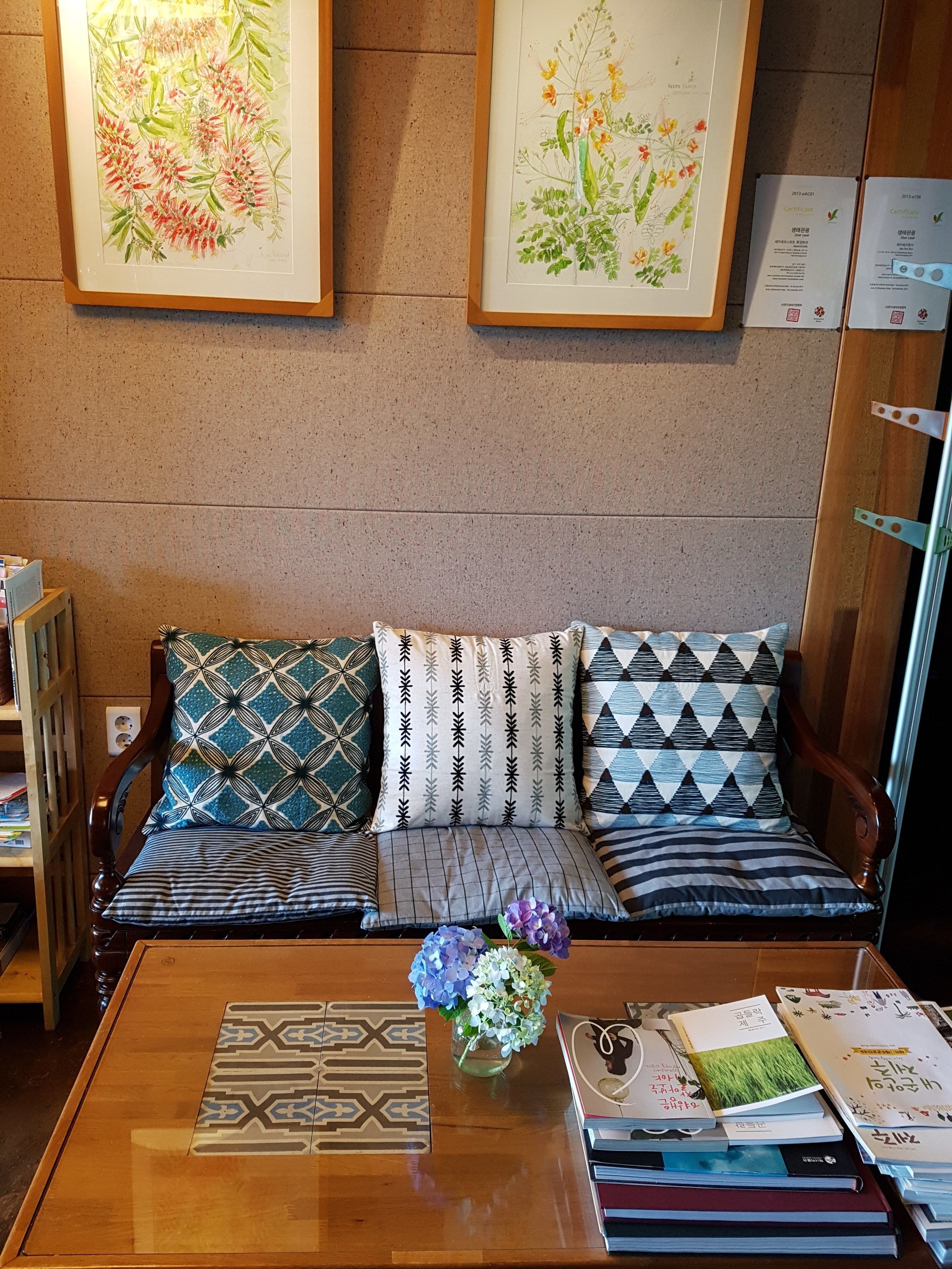 Ein gemütliches Wohnzimmer mit einem Holzsofa, gemusterten Kissen, einem Couchtisch mit Büchern und einer Blumenvase, sowie gerahmten Pflanzenabbildungen an der Wand.