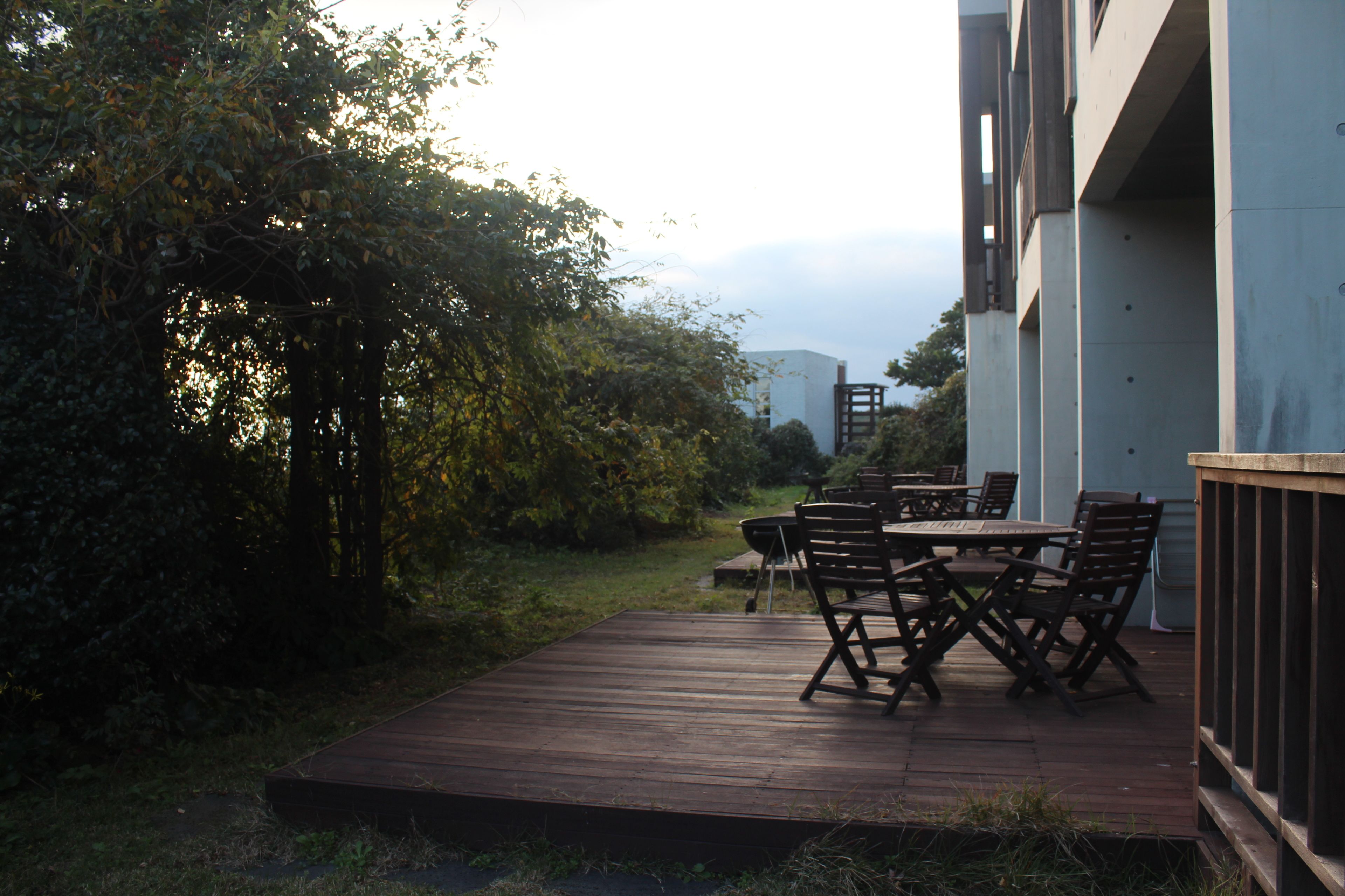 A wooden deck with outdoor furniture, surrounded by greenery and a building in the background.