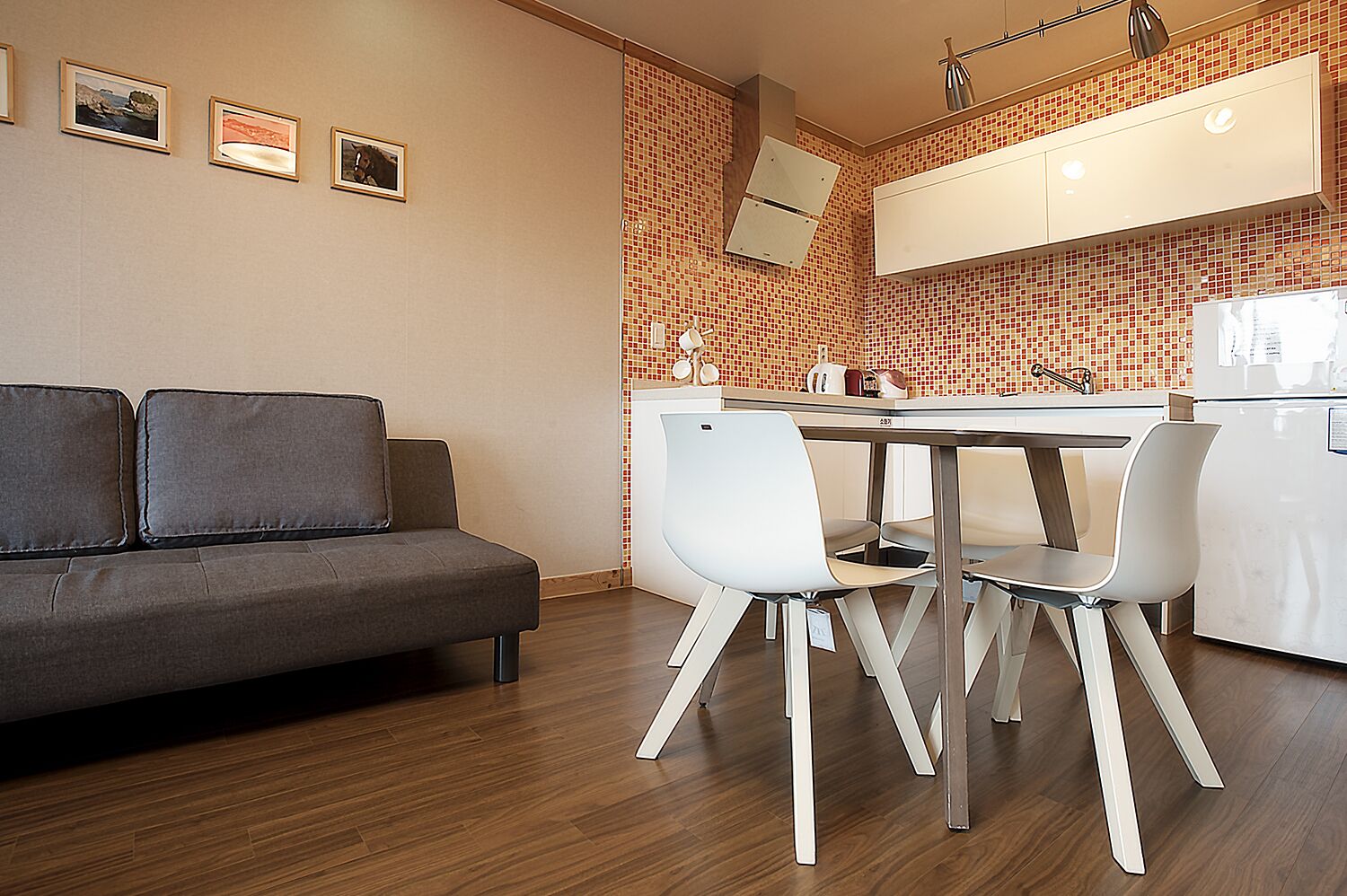A modern kitchen with a dining area, featuring a grey sofa, white chairs, and a tiled backsplash.