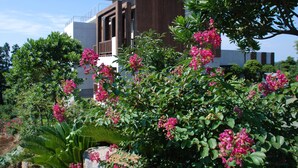 Un edificio moderno con balcón, rodeado de vegetación exuberante y plantas florecientes de color rosa.