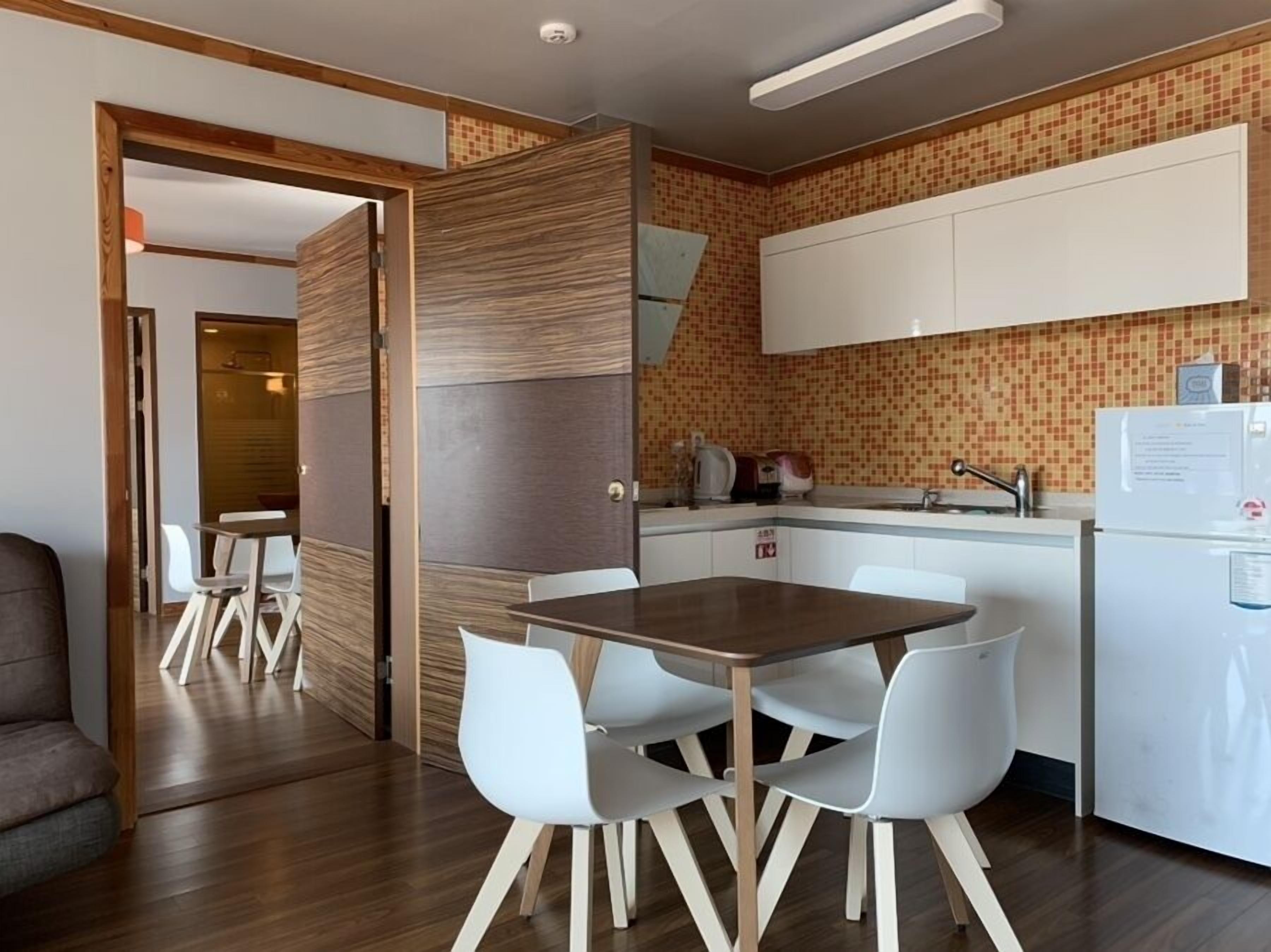 A compact kitchen with a dining area, featuring a wooden table and white chairs, a white refrigerator, and a tiled backsplash.