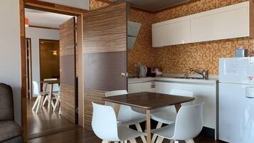 A compact kitchen with a dining area, featuring a wooden table and white chairs, a white refrigerator, and a tiled backsplash.
