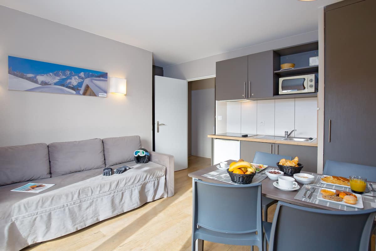 A compact hotel room with a sofa, dining table, breakfast setup, and a kitchenette.