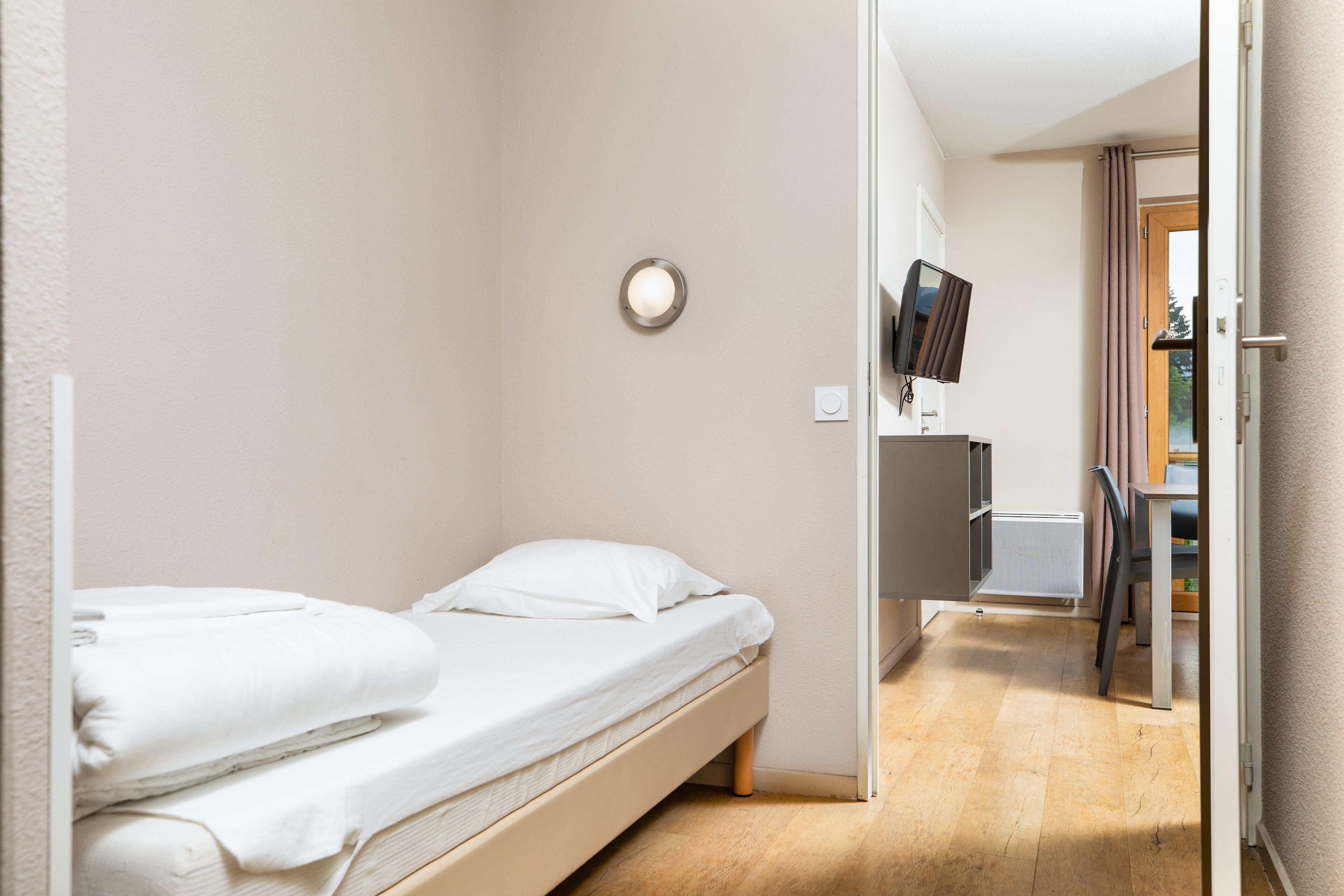 A small, clean room with a single bed, a flat-screen TV mounted on the wall, and a wooden floor.