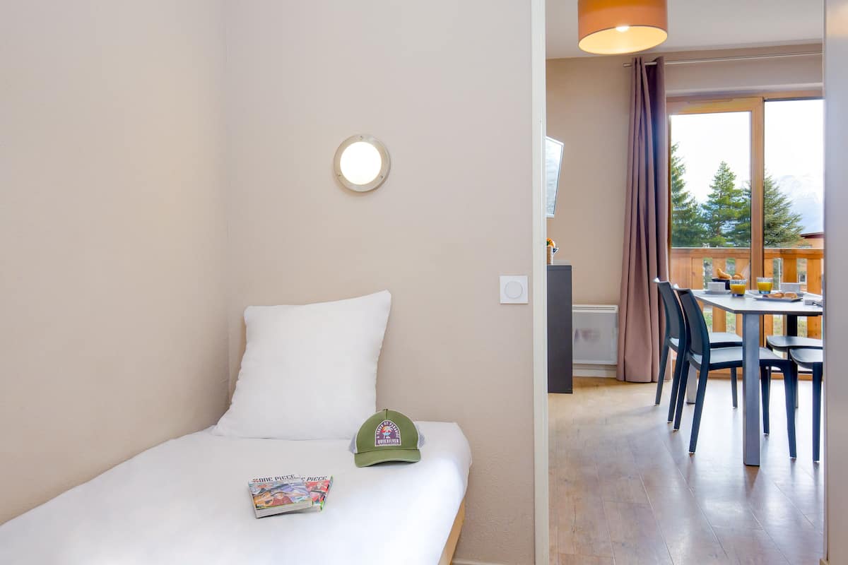 A hotel room with a bed, a small table with chairs, a window with a view of trees, and a ceiling light.