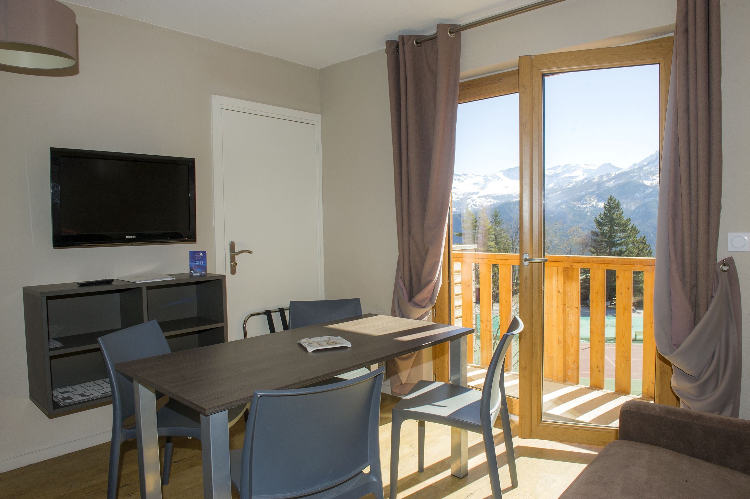 A room with a dining table, chairs, a TV mounted on the wall, and a balcony with a view of mountains.