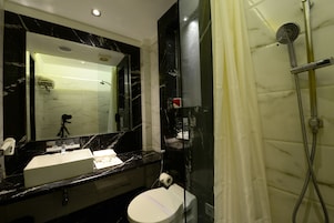 Une salle de bain avec un lavabo en marbre noir, des toilettes, une douche avec une paroi en verre et un miroir équipé d’une caméra montée sur un trépied.