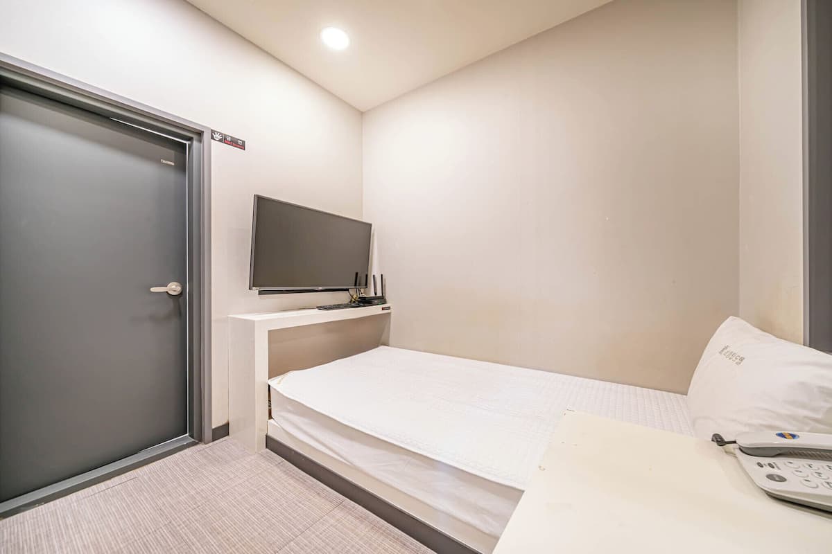 Una habitación pequeña y ordenada con cama, escritorio con teléfono y televisor montado en la pared, además de una puerta.