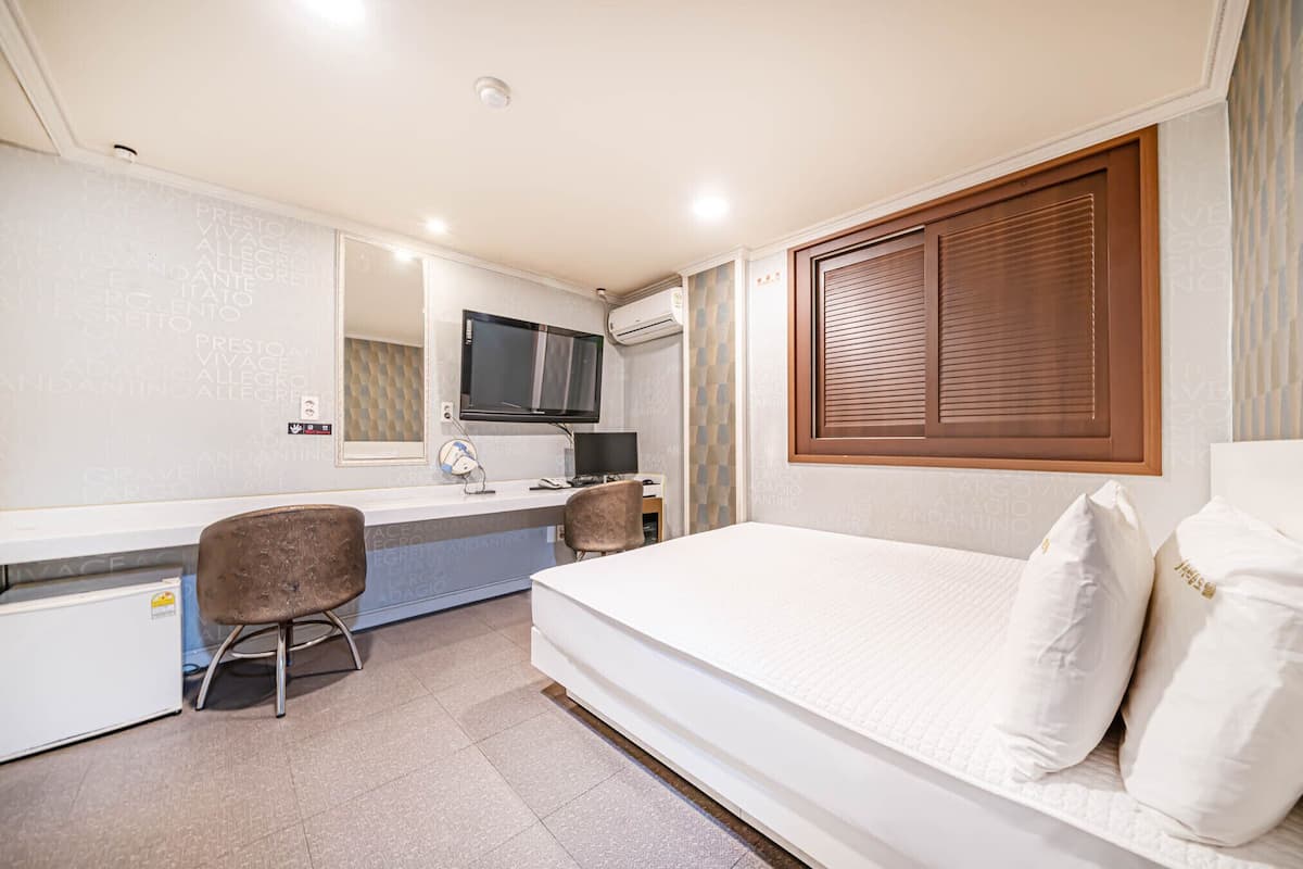 Una habitación de hotel con cama, escritorio con silla, televisión y ventana con persianas.