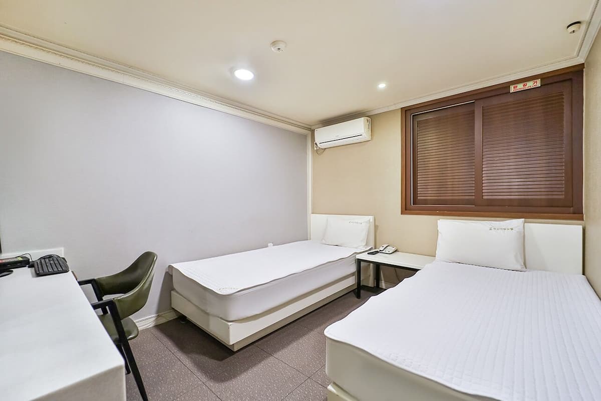 Habitación de hotel con dos camas individuales, un escritorio con silla, una mesita y un equipo de aire acondicionado.