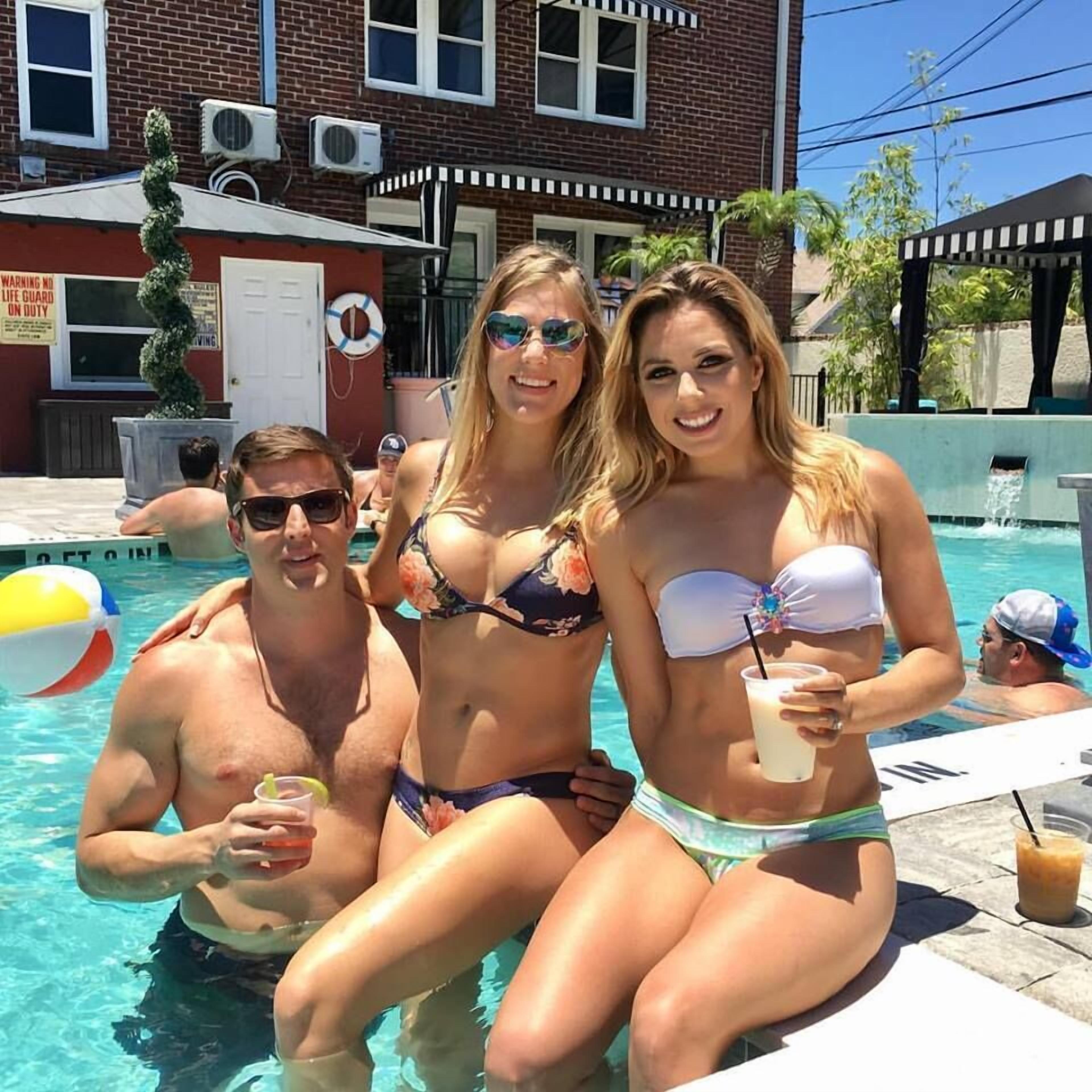 Uma área à beira da piscina com um edifício de tijolos ao fundo, um posto de salva-vidas e uma palmeira.
