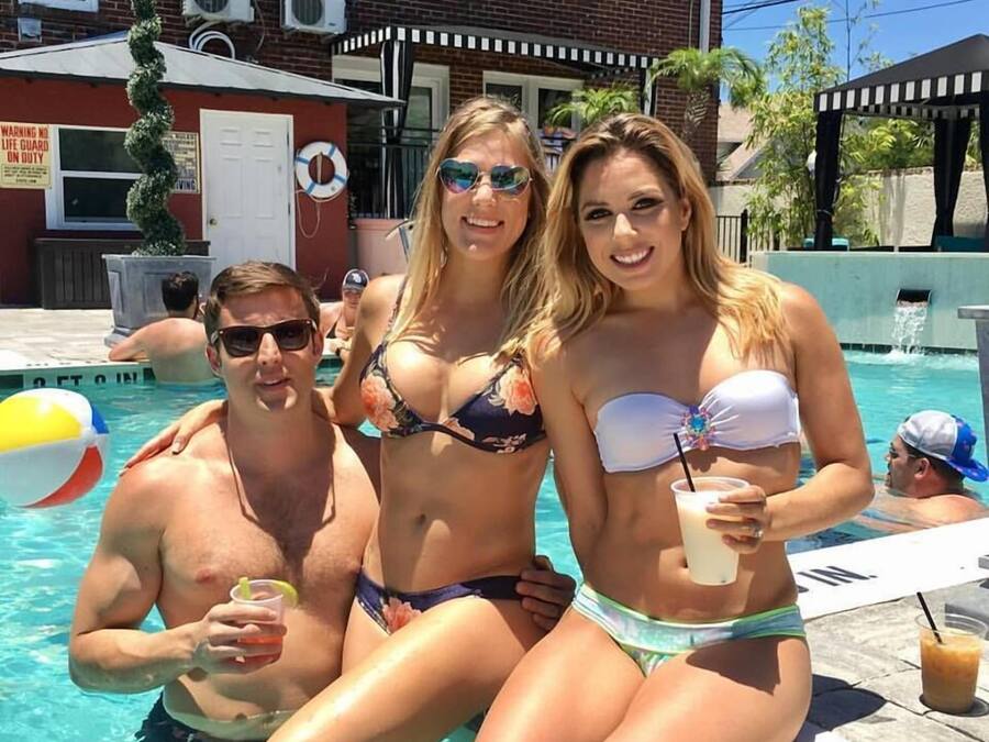 Uma área à beira da piscina com um edifício de tijolos ao fundo, um posto de salva-vidas e uma palmeira.