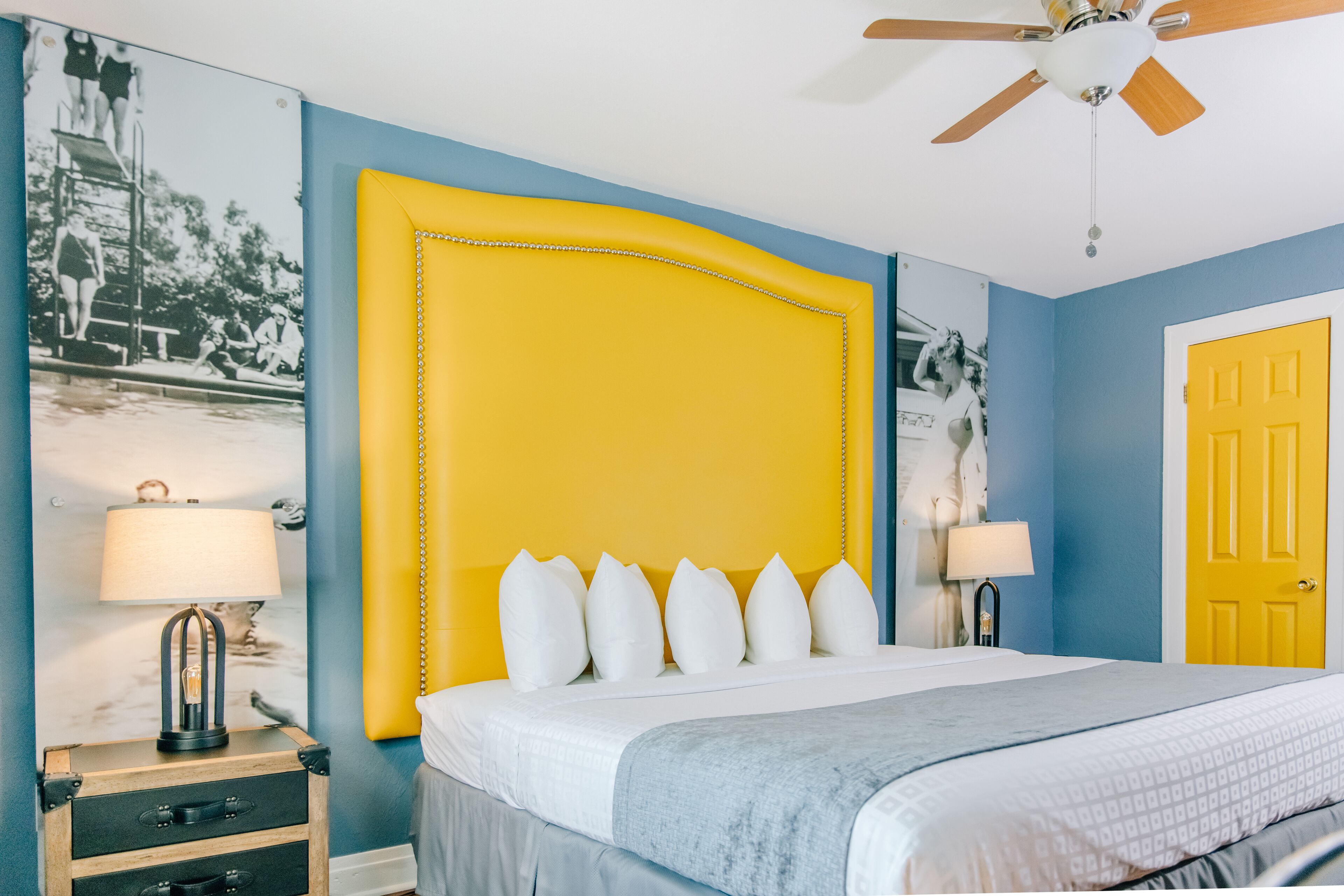A bedroom with a large bed, two bedside tables with lamps, a ceiling fan, and a yellow door.