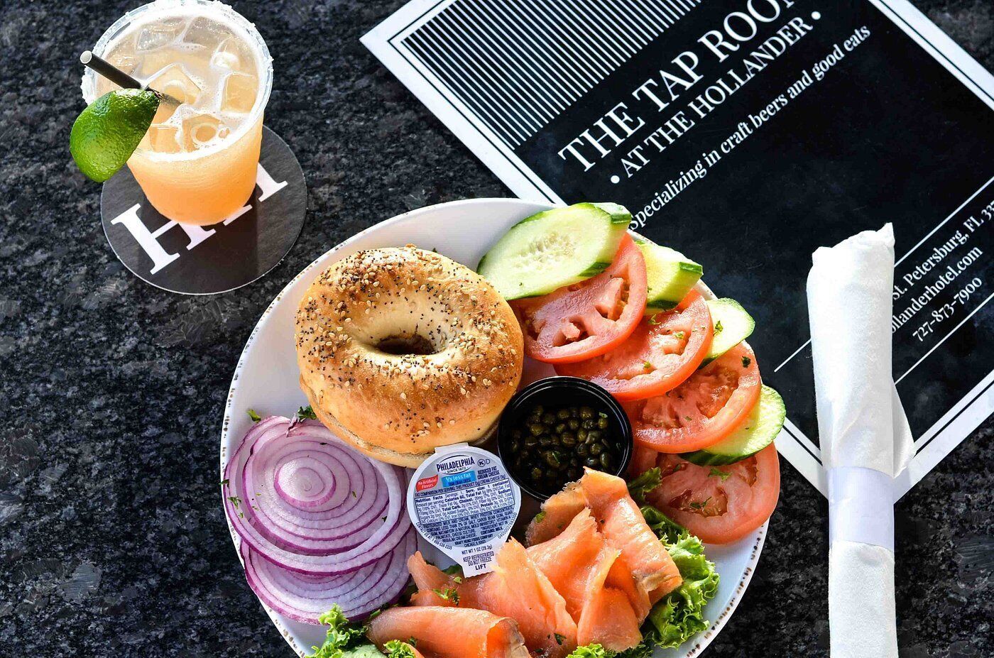 Ein Teller mit einem Bagel, Räucherlachs, Gemüse und einem Getränk, zusammen mit einer Speisekarte und einem Servietttuch.