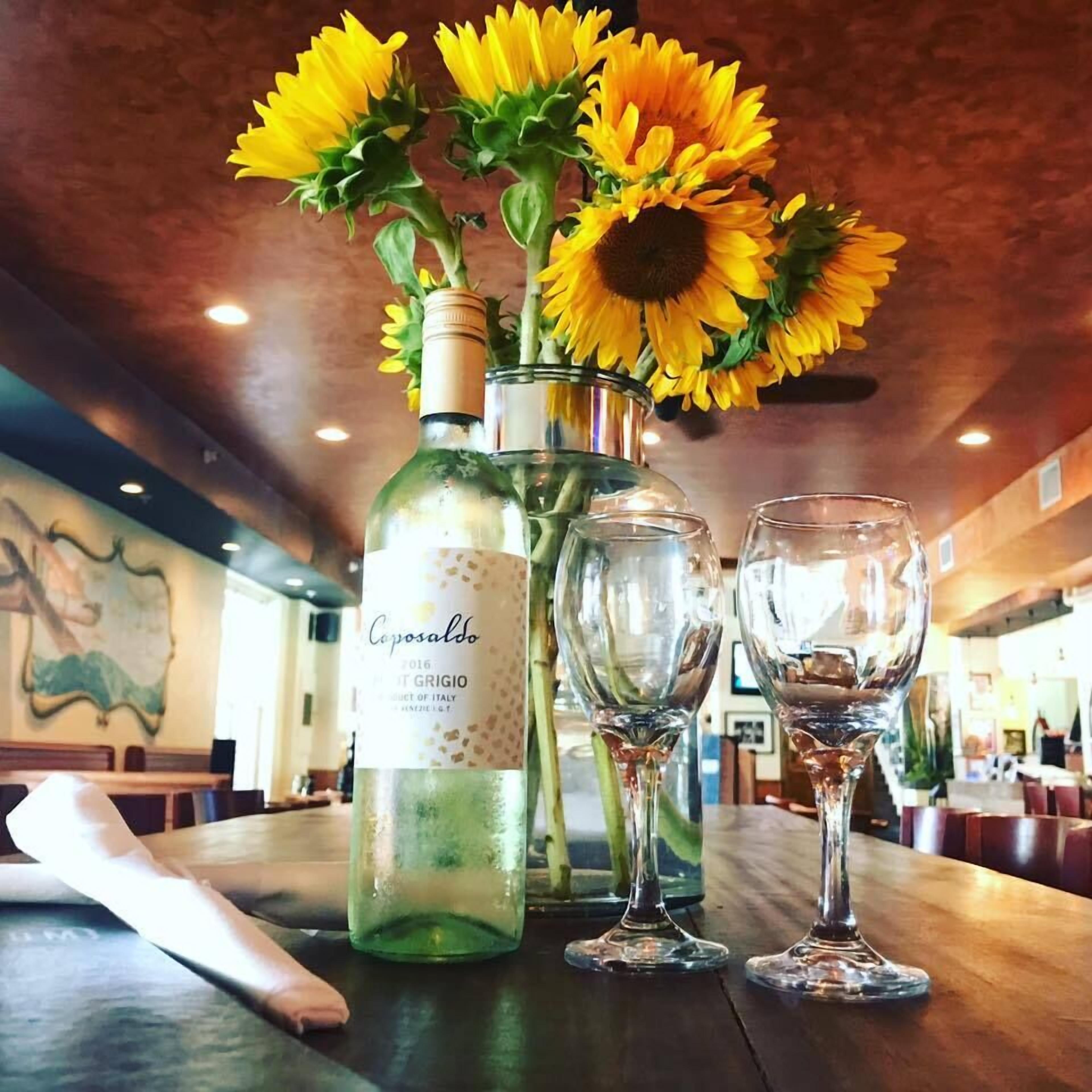 A table with a bottle of wine, two wine glasses, and a vase with sunflowers.