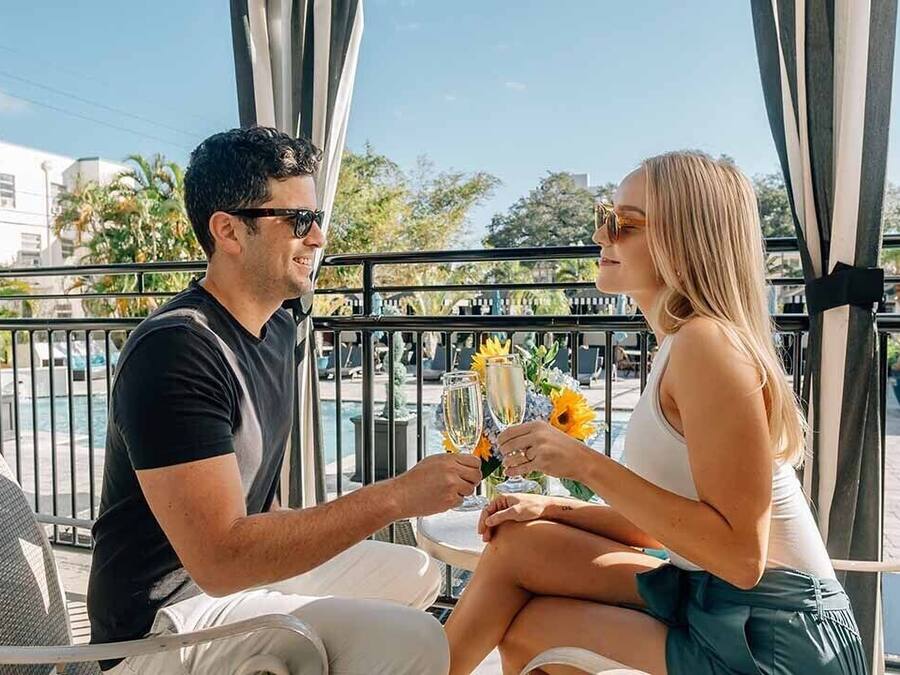 A couple toasting with champagne glasses on a balcony with a pool view.