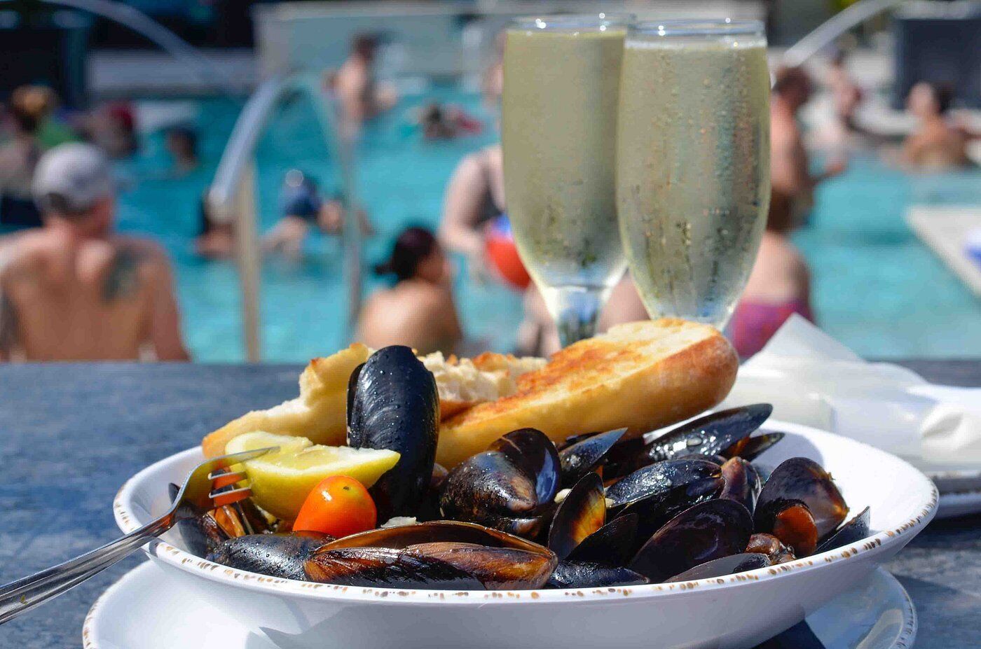 Un plato de mariscos con mejillones y pan, una copa de vino blanco, y una piscina al fondo.