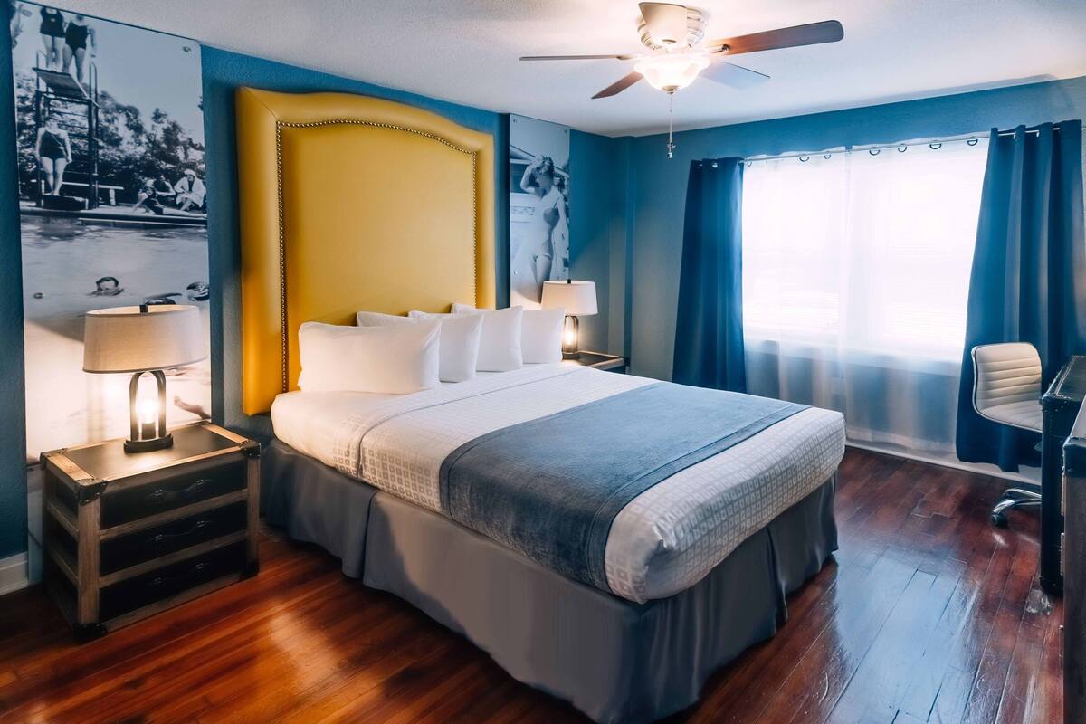 A hotel room with a large bed, a ceiling fan, a desk, and a window with curtains.