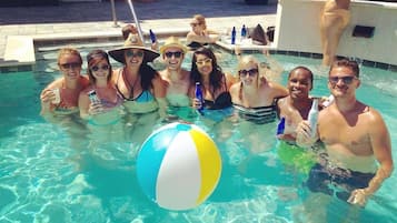 A group of people in a swimming pool with a beach ball, lounge chairs, and a poolside area.