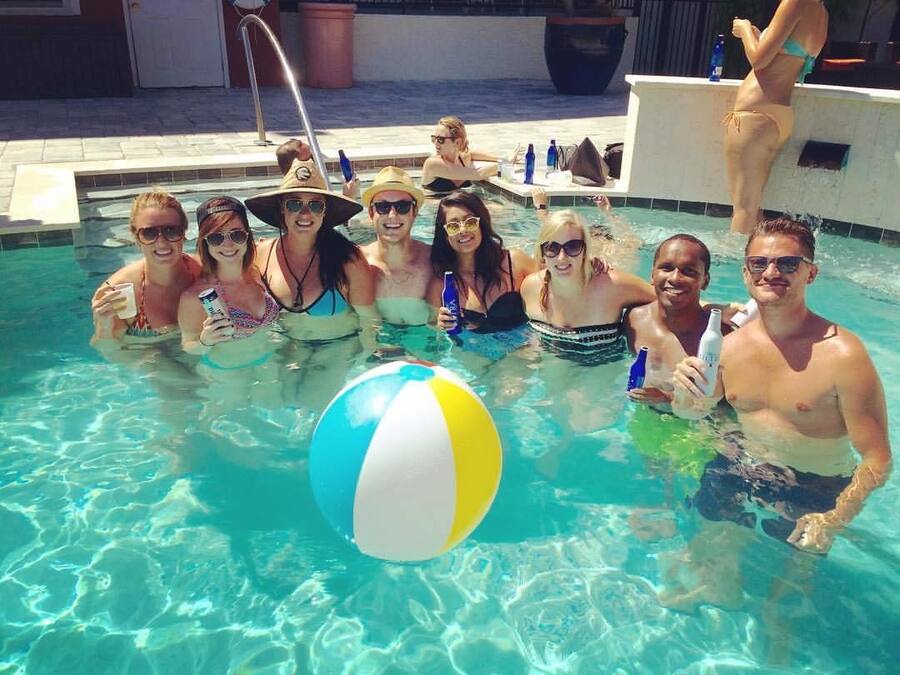 A group of people in a swimming pool with a beach ball, lounge chairs, and a poolside area.