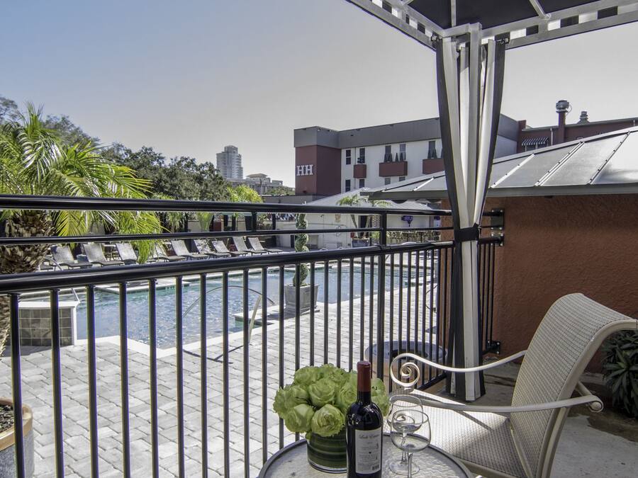 Uma varanda com piscina, cadeiras de descanso, uma mesa com garrafa e copo, e vista para edifícios e árvores.