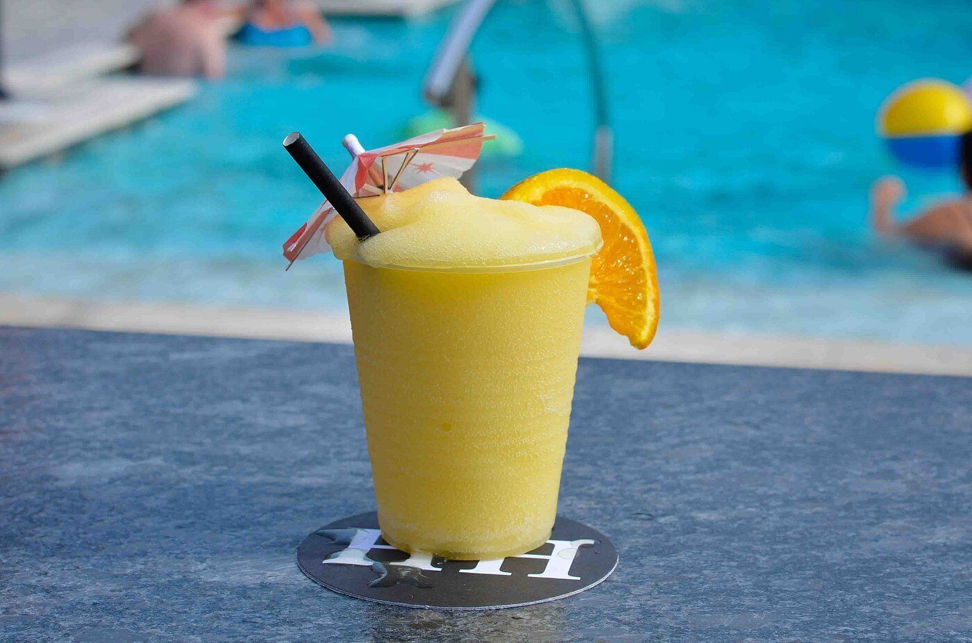 Un vaso alto de una bebida amarilla, con pajita y una rodaja de naranja en el borde, sobre un posavasos cerca de una piscina.