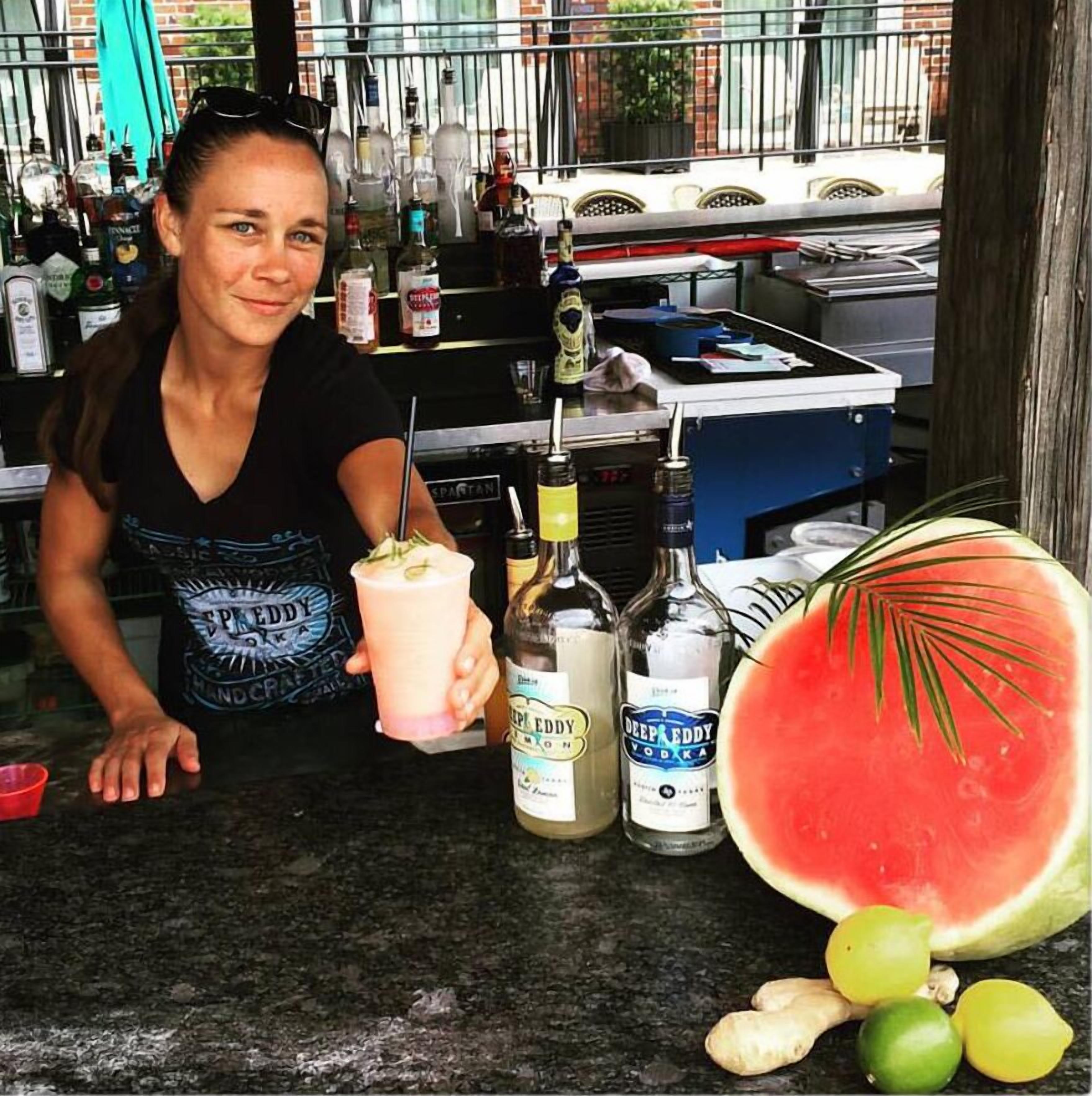 A bar counter with a woman holding a drink, bottles of Deep Eddy Vodka, a watermelon, and some limes.