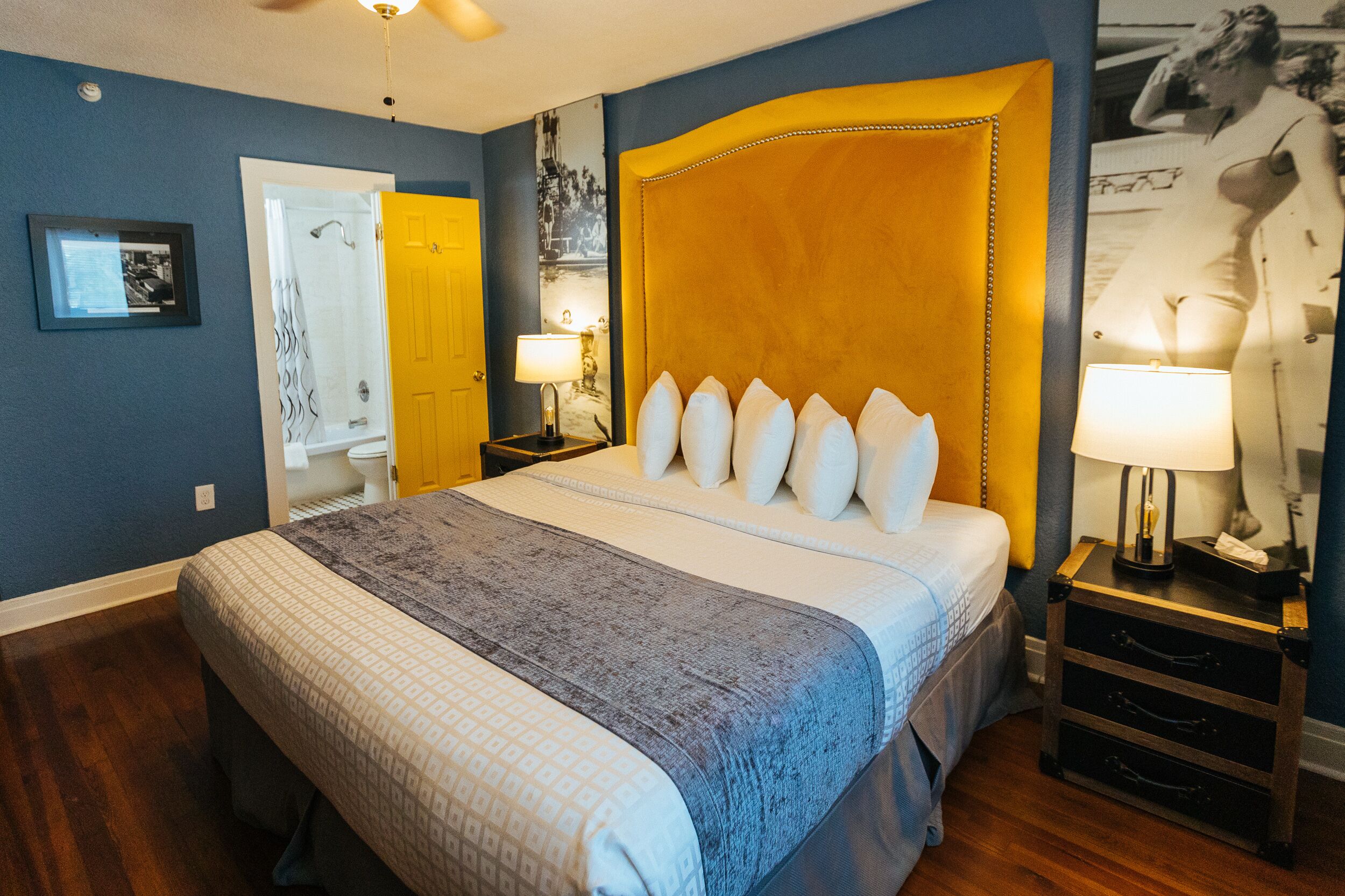 A bedroom with a large bed, a nightstand with a lamp, a door, and a framed picture on the wall.