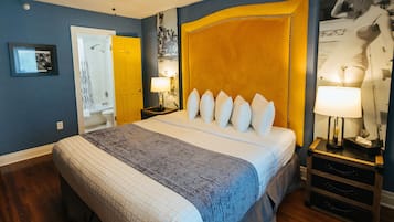 A bedroom with a large bed, a nightstand with a lamp, a door, and a framed picture on the wall.